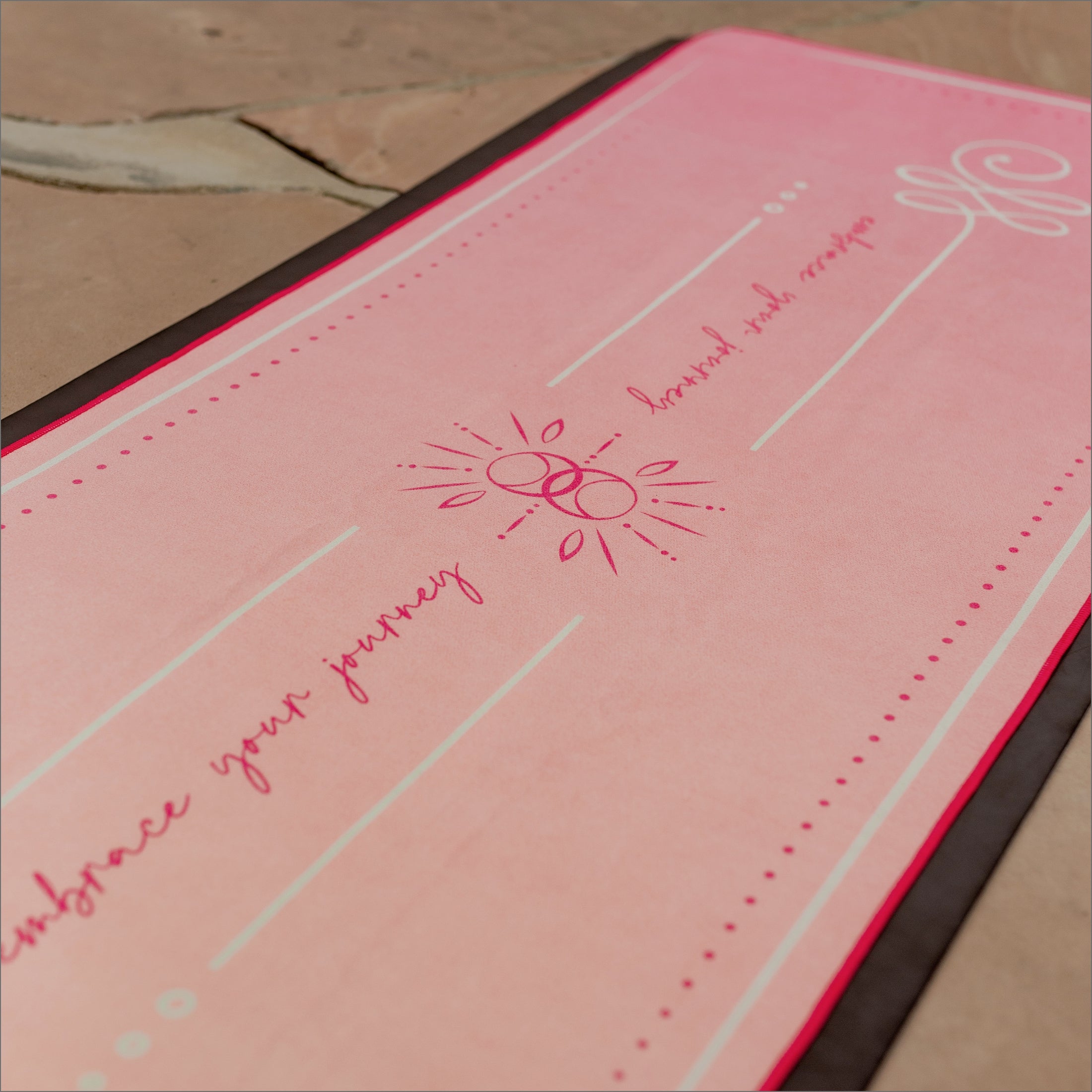 Close-up of Glow towel’s sun-fleur motif on coral-pink microfiber — accented with delicate script ‘embrace your journey’, showing textured fabric grip.