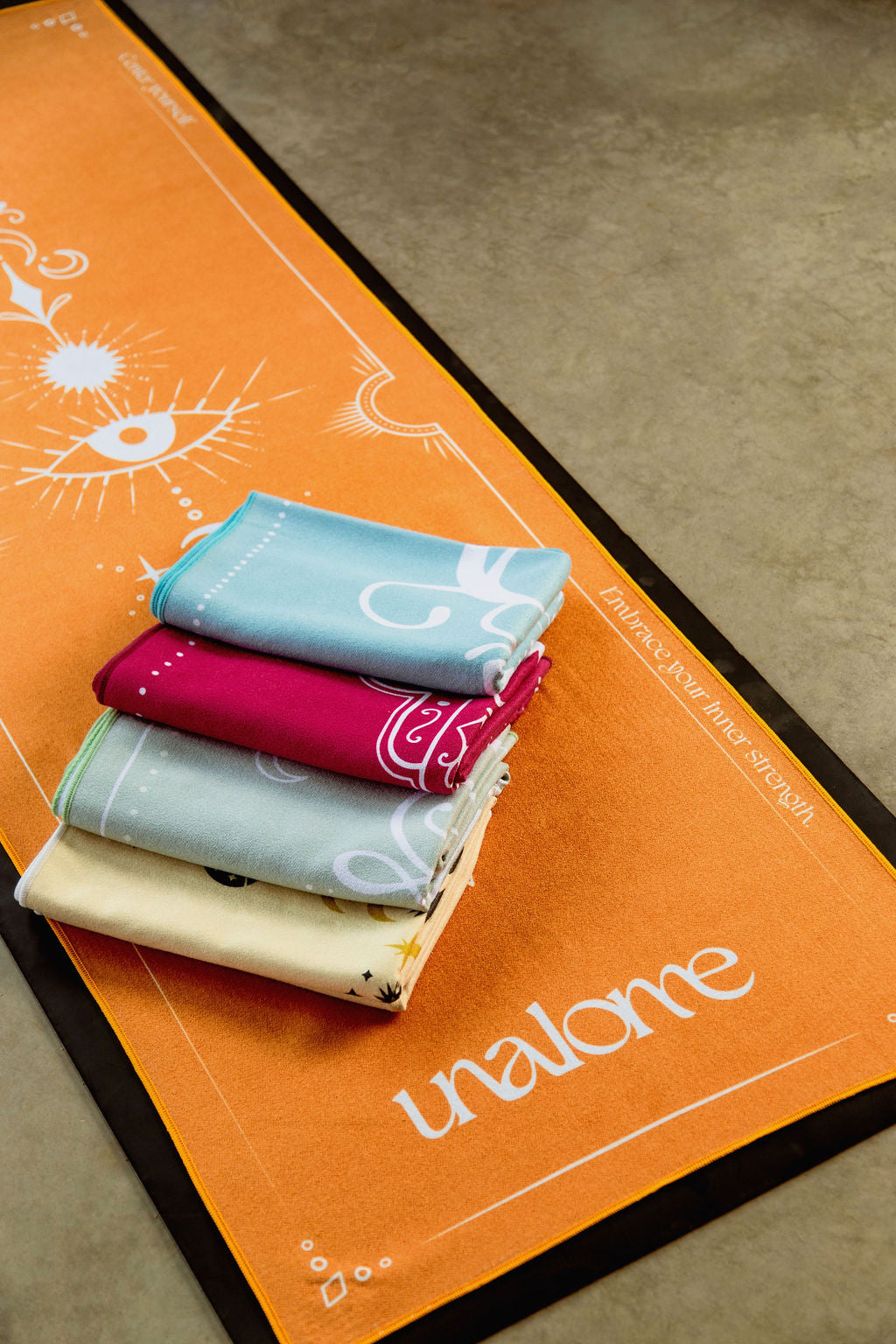 Stack of folded Grounded Collection from Unalome Yoga Hot Yoga Towel collection; orange, blue, yellow, green and pink towels