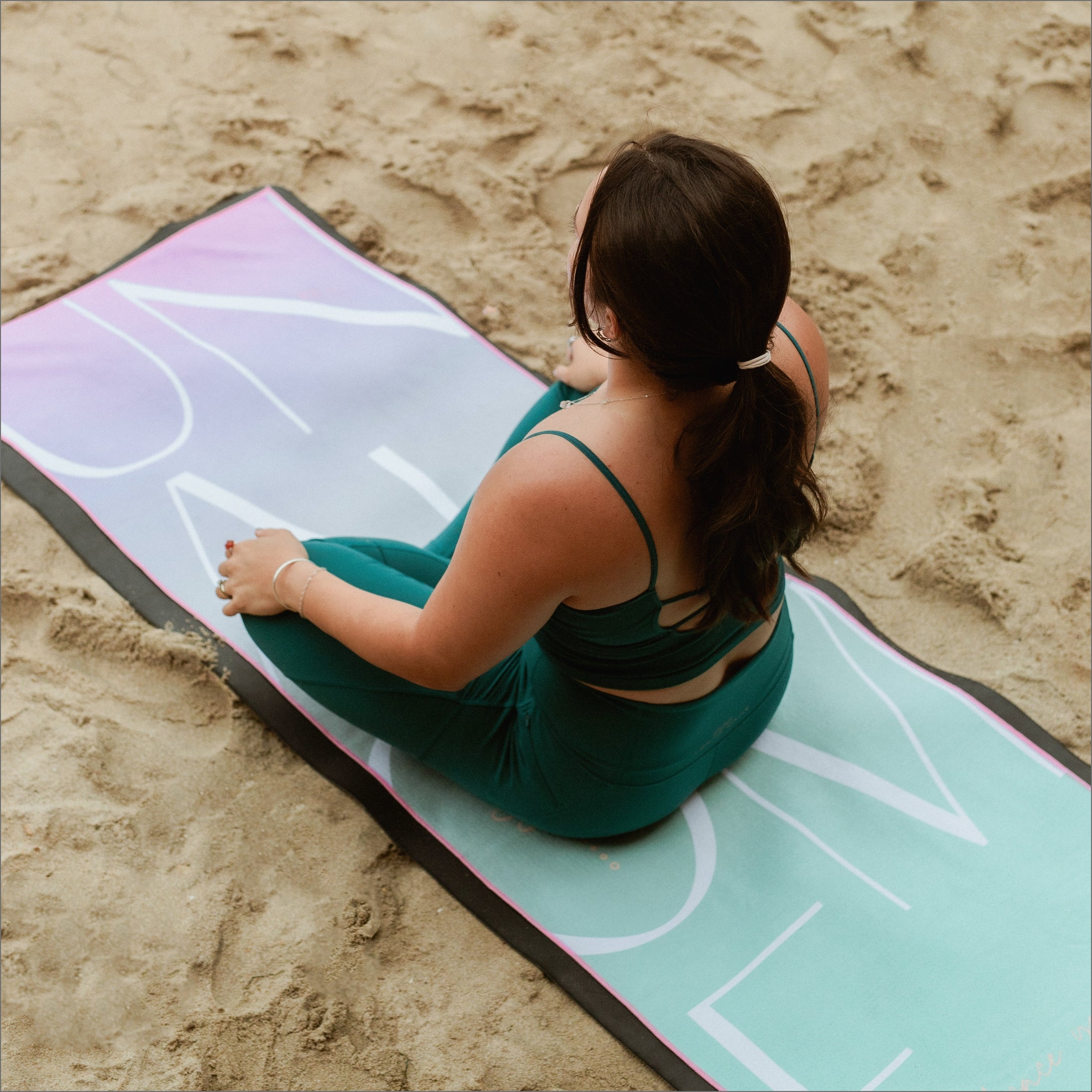 Woman practicing yoga on Unalome Santi towel at the beach — pastel gradient towel with soft mint and lavender tones, bold white minimalist lettering, and coral pink trim, designed for hot yoga and vinyasa flow.